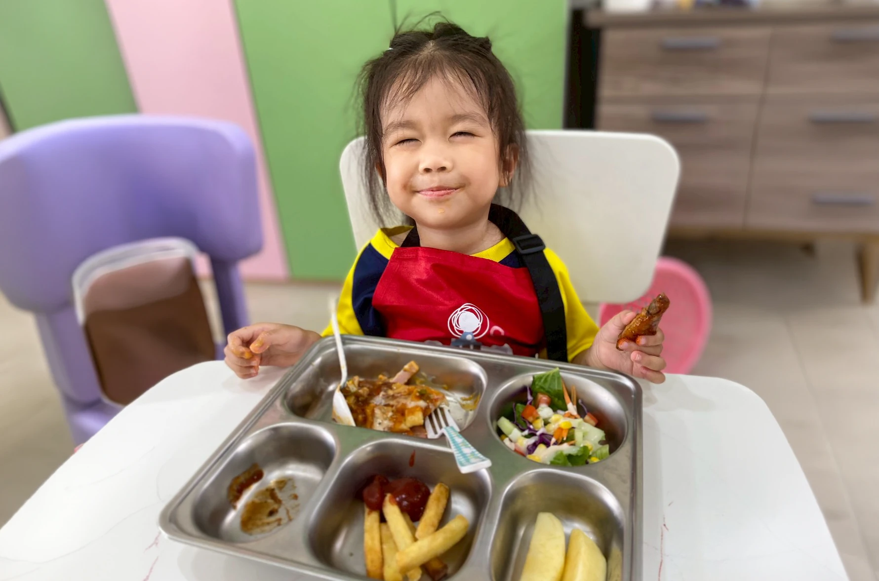 A child eating and smiling with her meal tray of chicken, fries, salad and fruit in front of her