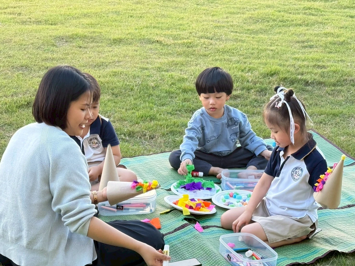 Children sitting on the grass making paper crafts with their teacher