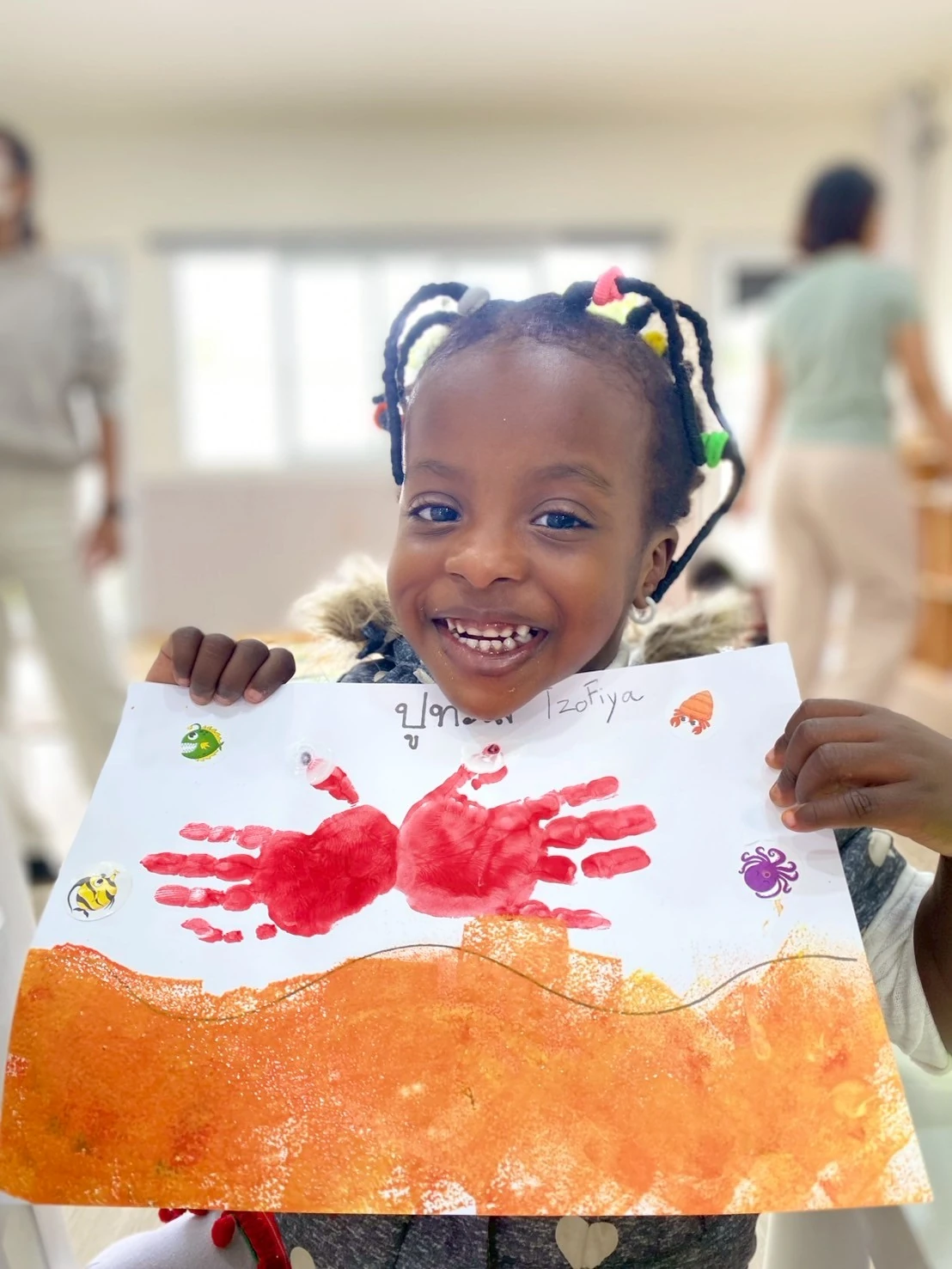 A Nursery child smiling while holding up a handprint painting she made