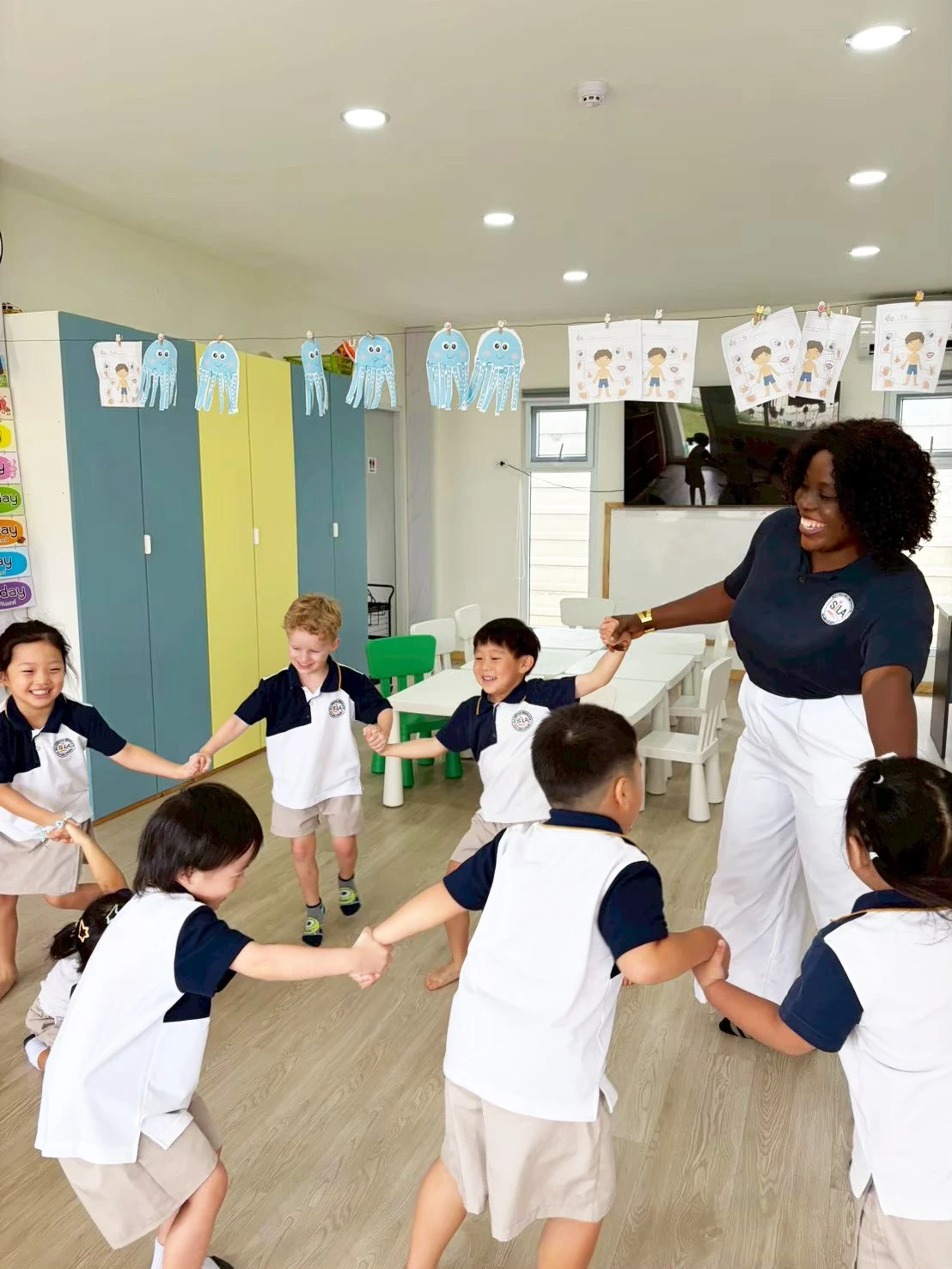 Children and their teacher holding hands in a wide circle game across the classroom floor