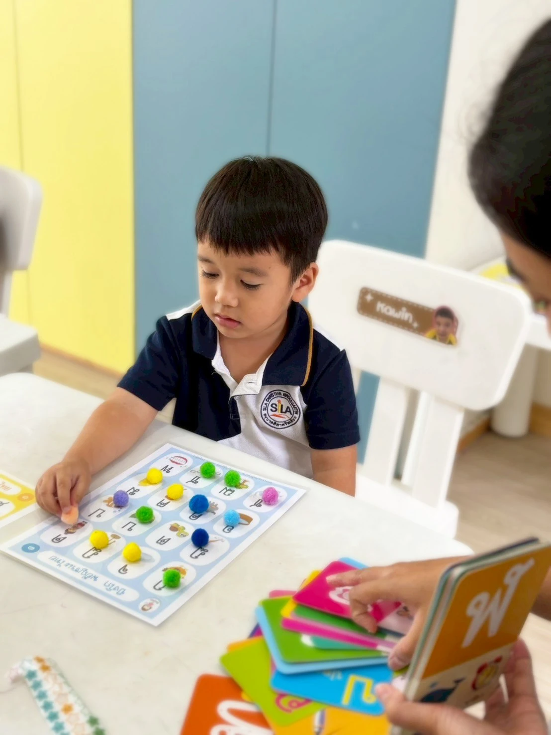 A child placing coloured pom-poms onto a Thai alphabet board while a teacher holds up Thai letter cards