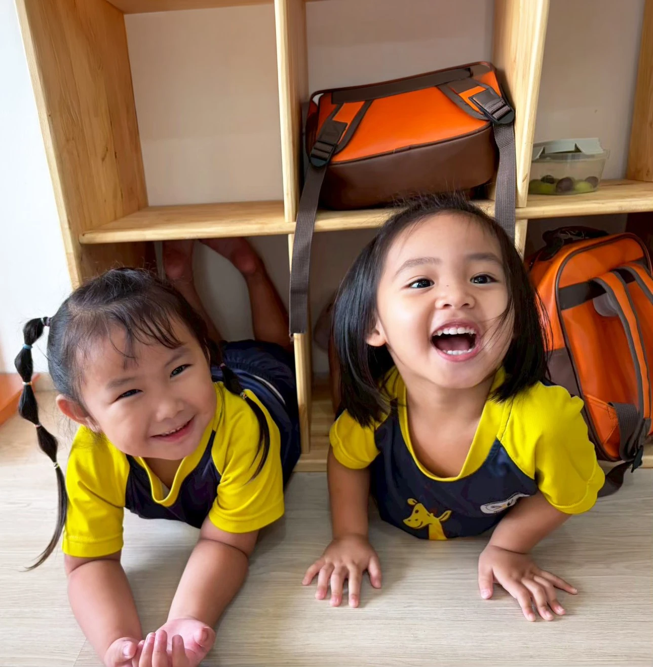 Two Kindergarten children lying on the classroom floor beneath the cubby shelves, laughing up at the camera