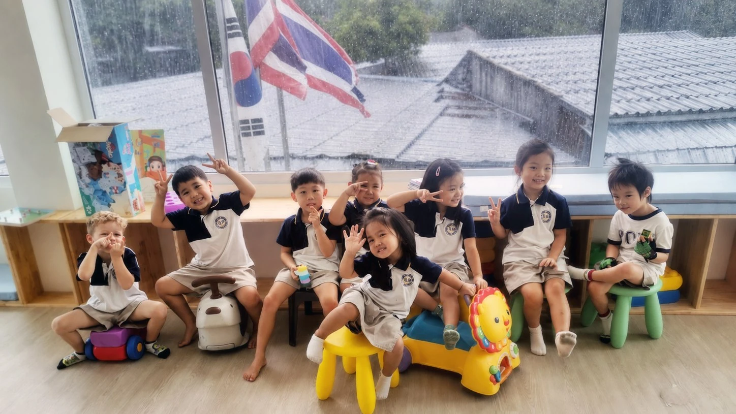 A group of Kindergarten children sitting together by the classroom window, smiling and waving at the camera