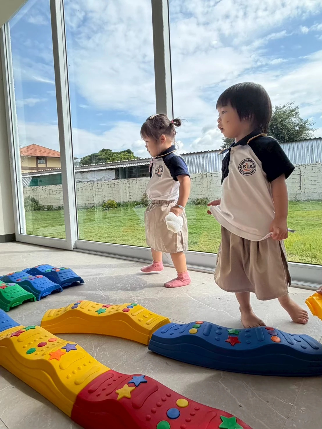 Two young children walking together across a sensory balance path in a bright, sunlit classroom
