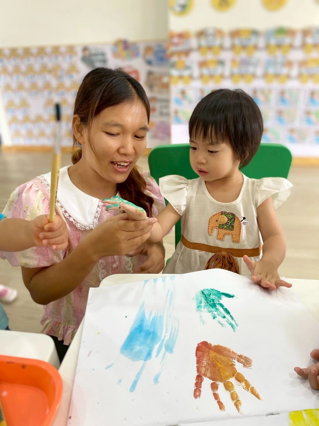 A teacher smiling warmly as a Nursery child watches her paint a handprint together