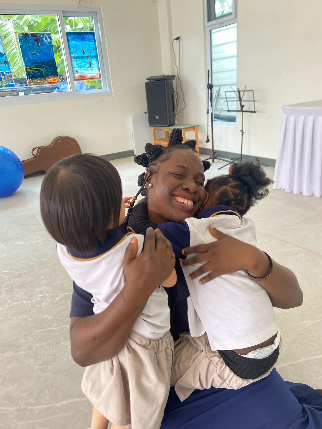 A teacher laughing warmly as two Nursery children hug her from either side