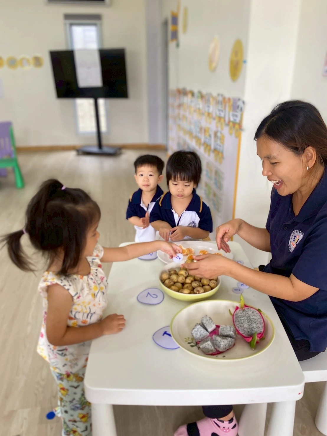 A teacher smiling as Nursery children gather around bowls of longan and dragon fruit at a table