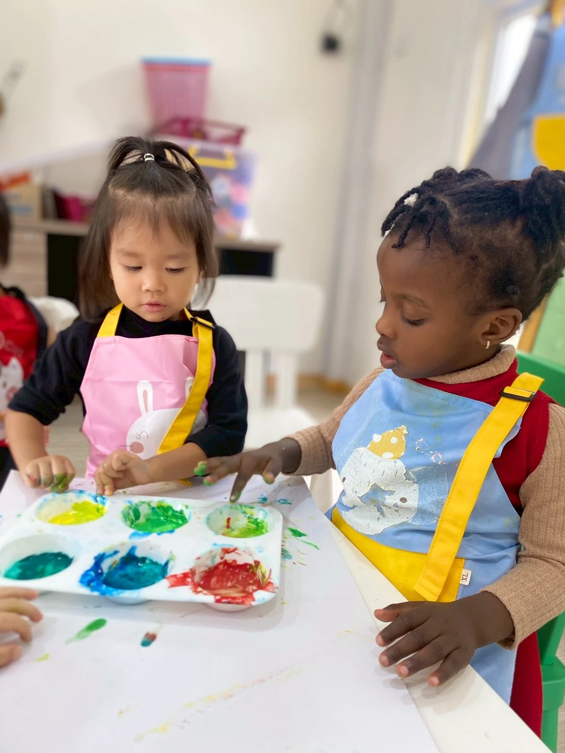 Two Nursery children in aprons finger-painting together at a table