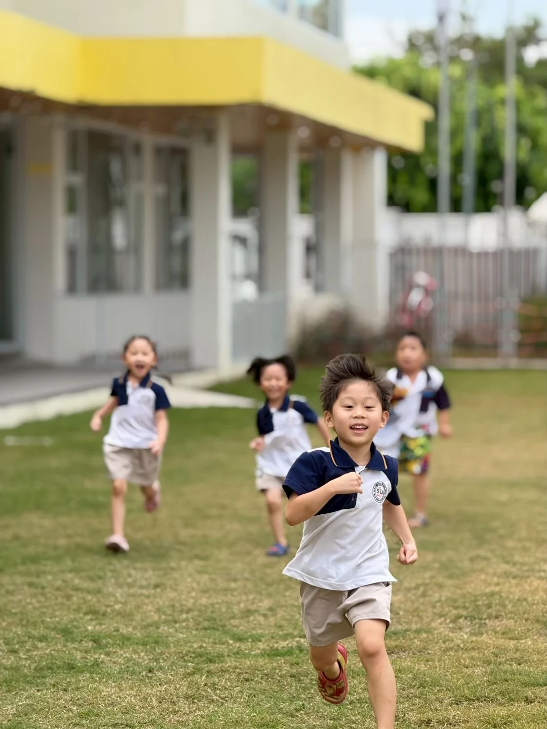 A SiLA boy running toward the camera across the school lawn with classmates behind him and the school building beyond