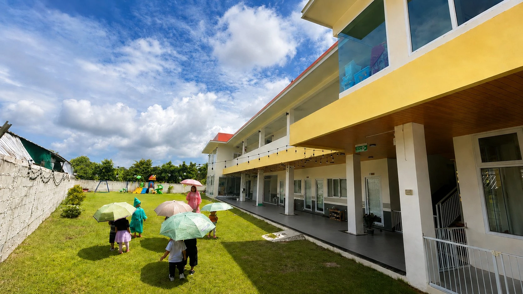 The SiLA school building and playground on a sunlit day, with children carrying colourful umbrellas crossing the lawn